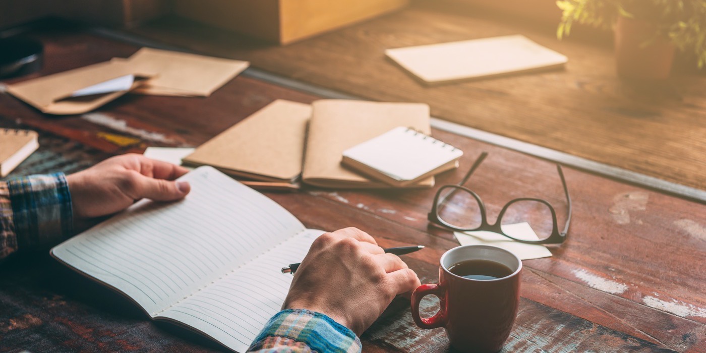 A notebook, a pen and a mug on a wooden desk in morning light.