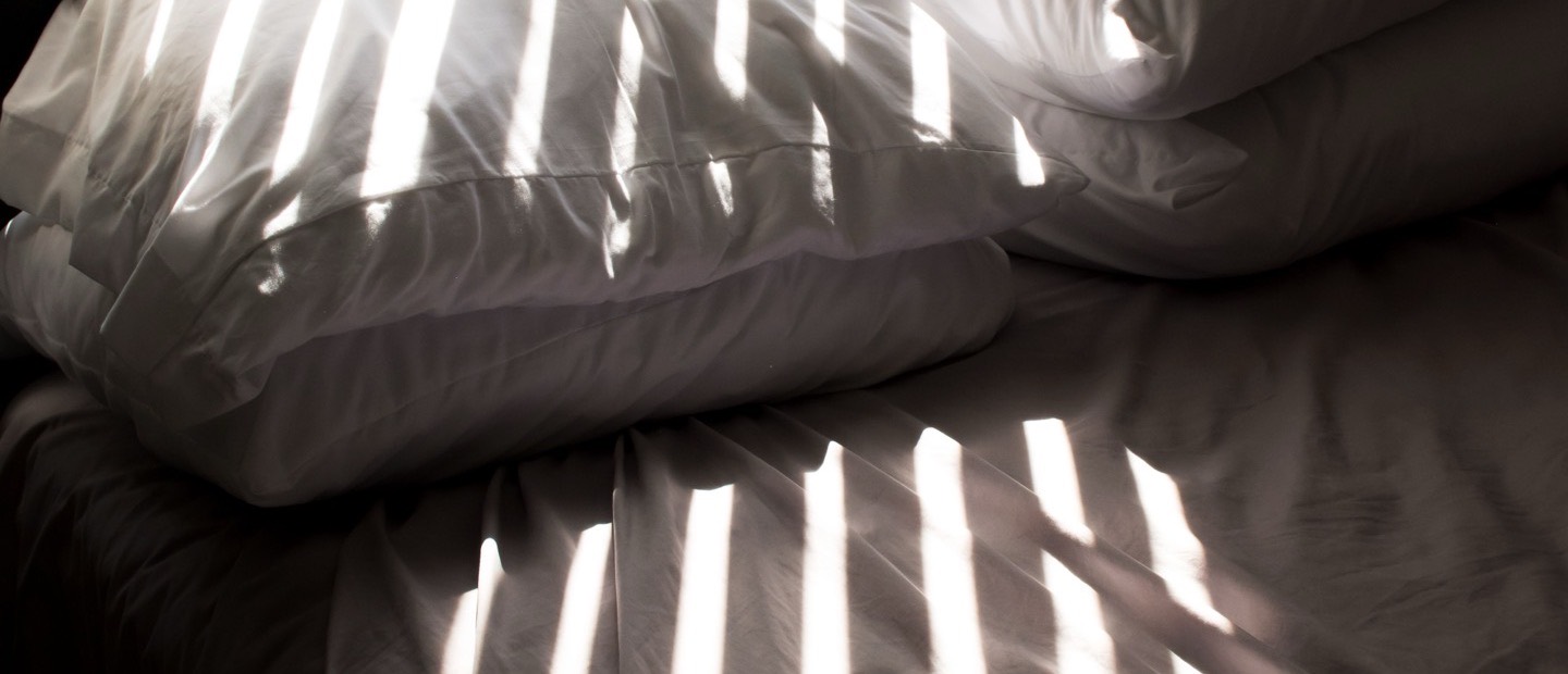 Bars of sunlight through a blind falling across a made bed.
