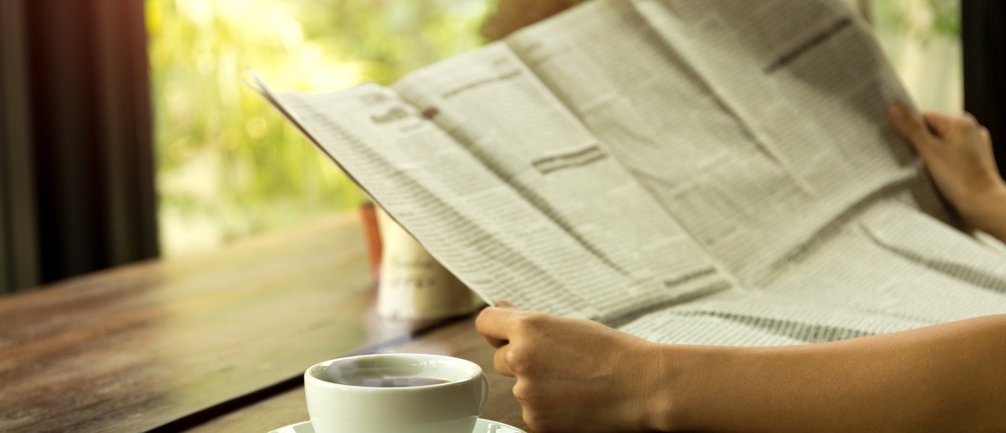 Hands holding an open newspaper at a cafe table, coffee beside it.